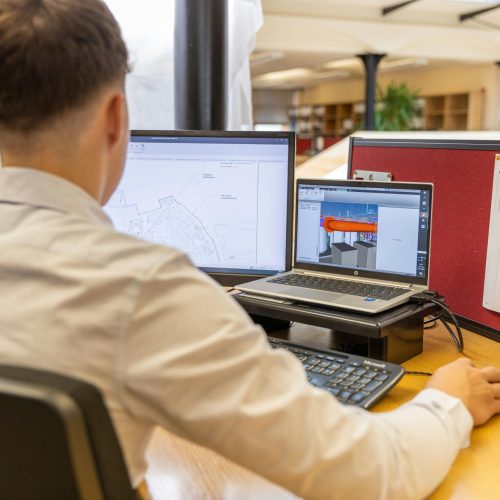 Back view of engineer focused on CAD design at dual monitors in bright office setting.
